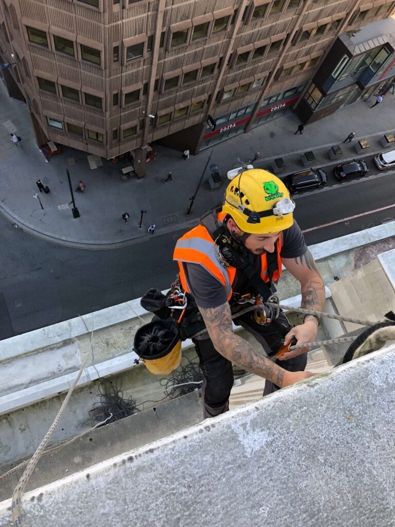 A man is performing inspections on the edge of a building, utilizing rope access for safe and efficient work.