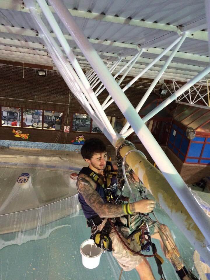 A man in a harness is painting a boat in an indoor pool.